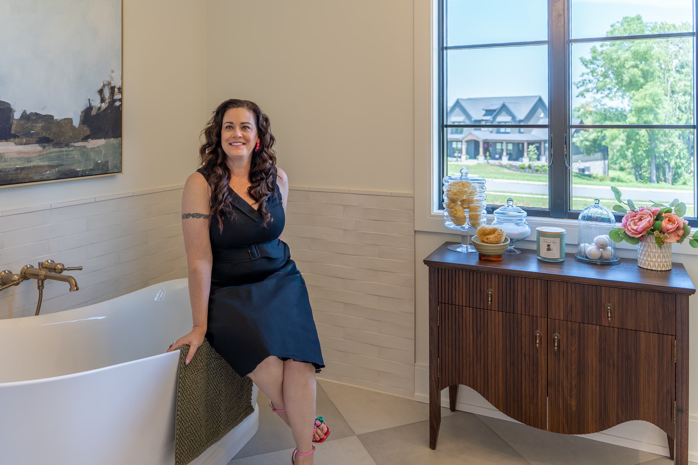 Shannon Clayton seated on the edge of a soaking tub in a finished bathroom, beside a walnut cabinet and a window