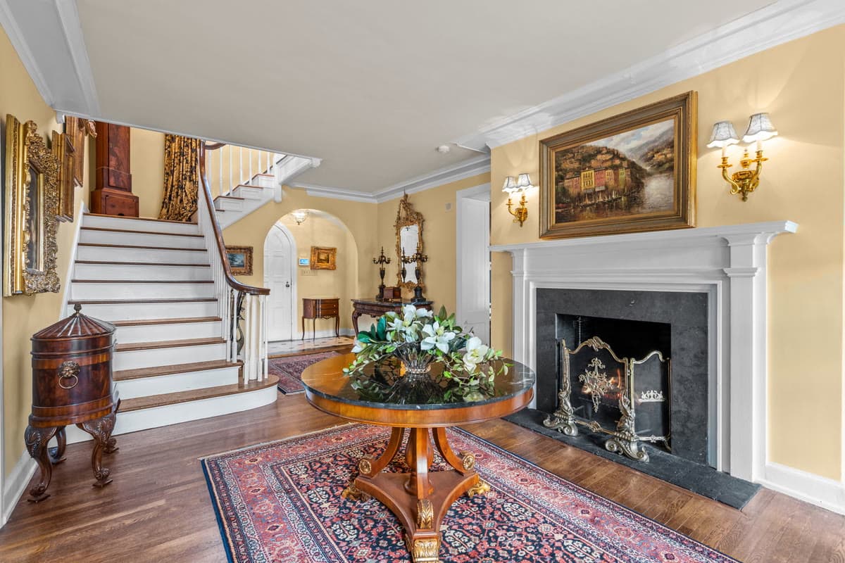 A front hall with a staircase, a round table of magnolias, and a marble fireplace beneath a gilt framed painting