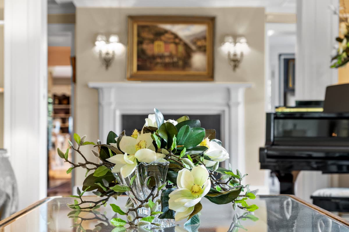 White magnolias in a crystal bowl on a mirrored table, with a fireplace and a grand piano behind