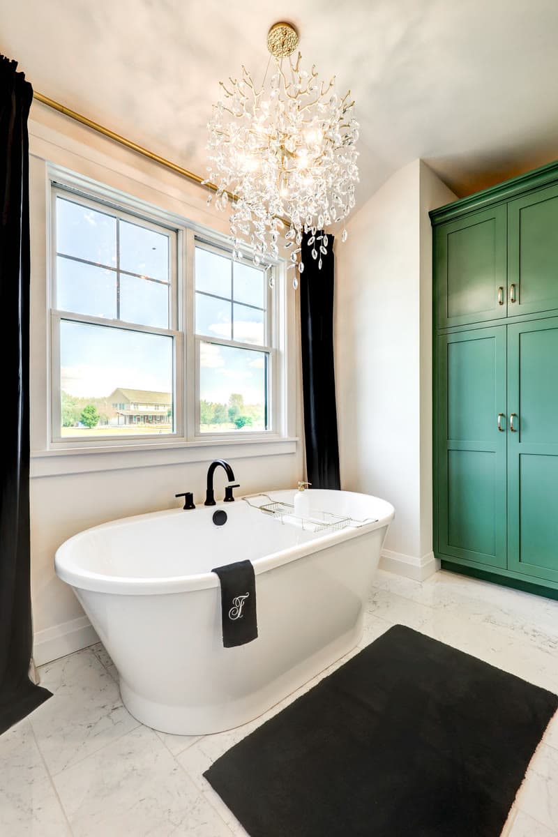 A freestanding tub beneath a crystal chandelier, beside a green linen cabinet