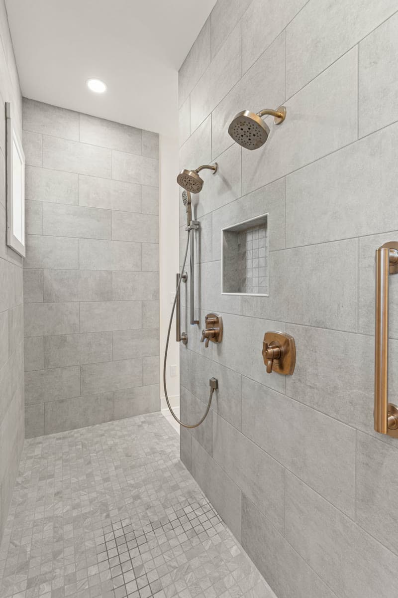 A curbless walk in shower in grey tile with brass fixtures and a grab bar